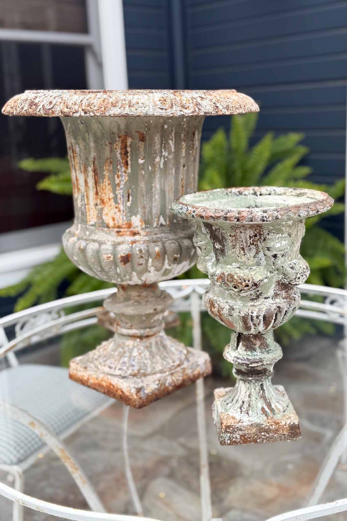Two ornate, weathered urn planters with chippy, distressed green and rust patina sit on a round glass table outdoors, with ferns and a blue house exterior in the background.