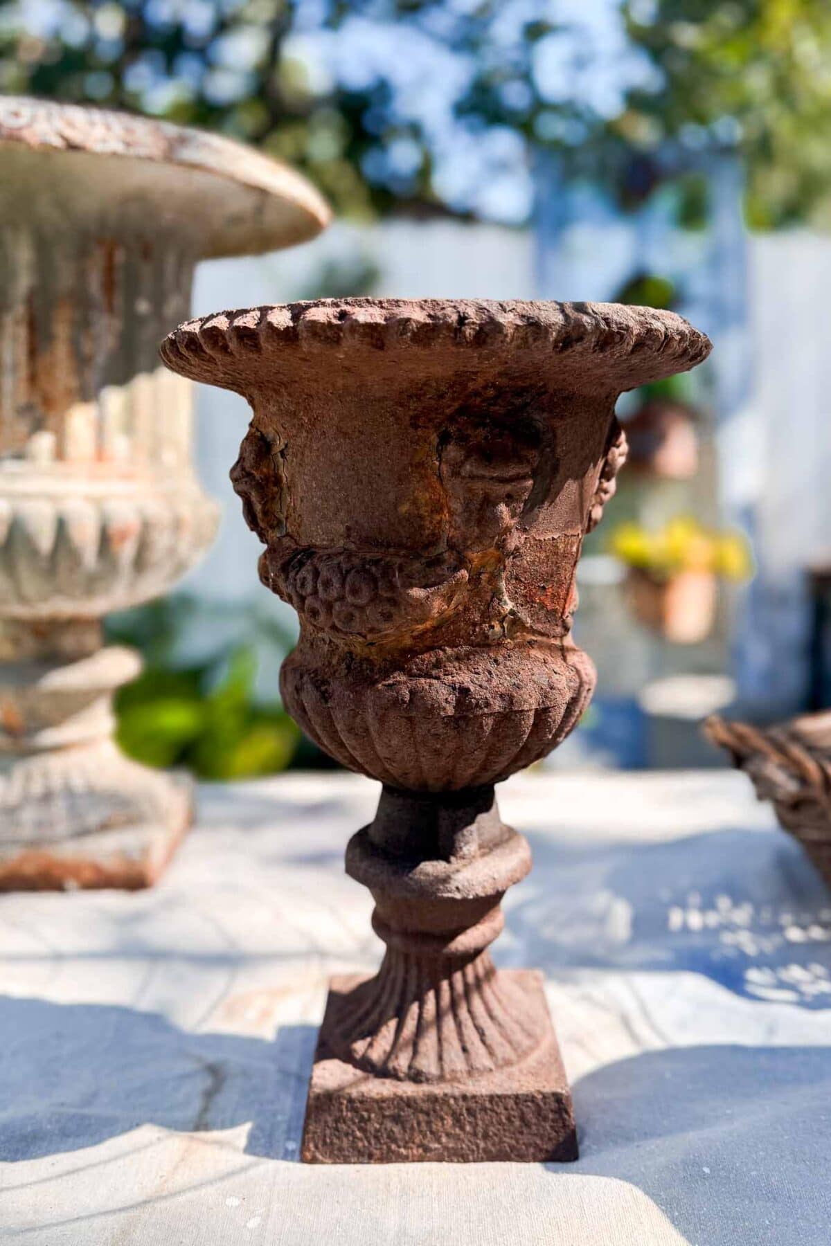 A weathered, ornate metal urn with vintage charm sits on a sunlit outdoor table, perfect for Spring entry table decor, with blurred greenery and another urn visible in the background.