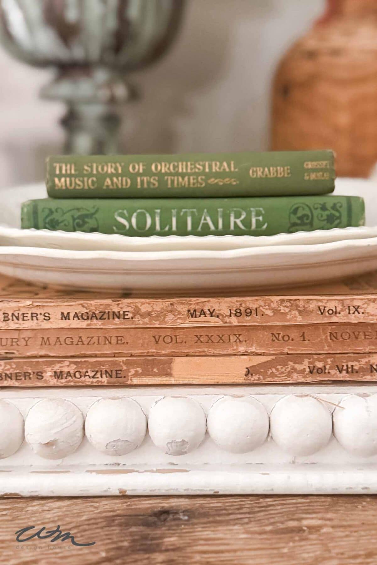 A stack of vintage books, including "THE STORY OF ORCHESTRAL MUSIC AND ITS TIMES" and "SOLITAIRE," brings vintage charm to a spring entry table decor, resting on a white tray above faded magazines with worn edges.