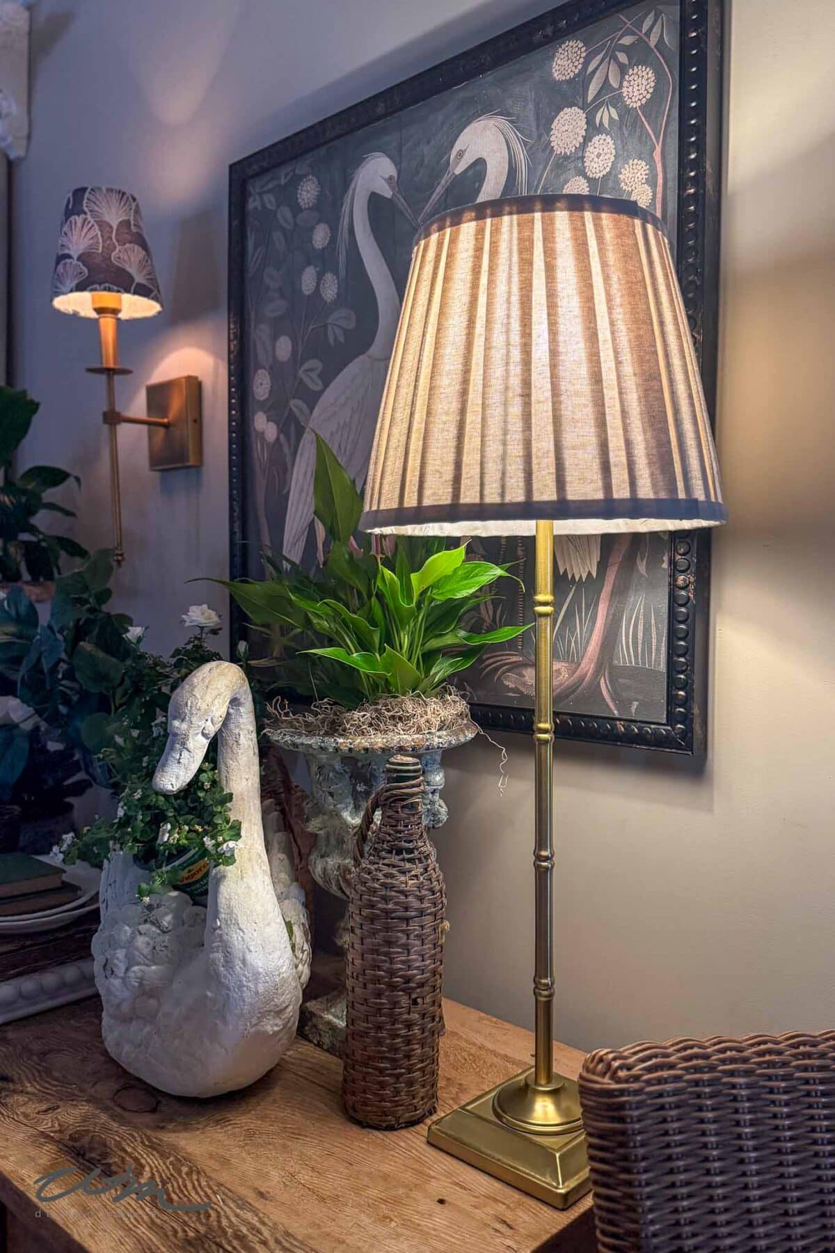 A cozy interior scene with a tall brass lamp on a wooden table, a decorative white swan, potted green plant, woven bottle, wall art featuring white birds, and a wall-mounted sconce in the background.