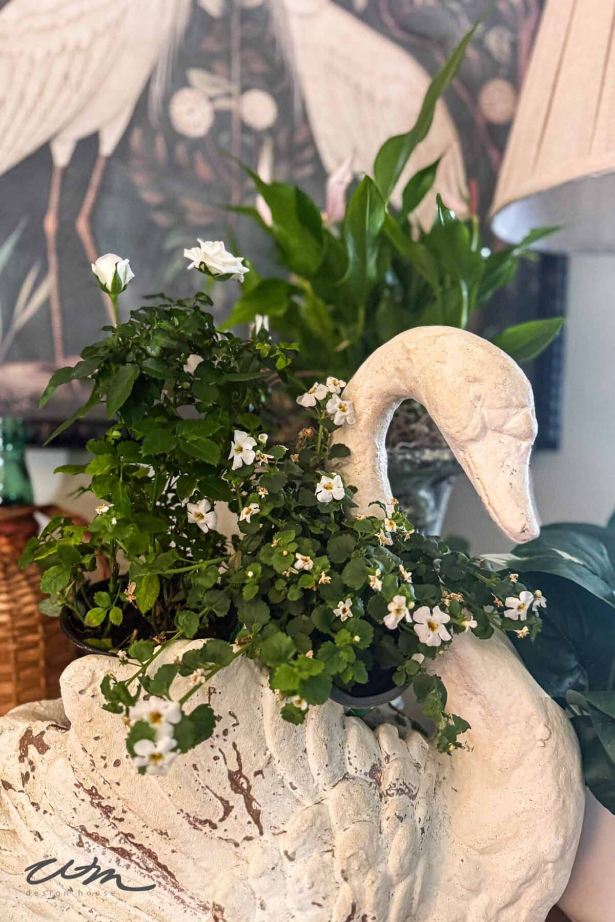 A white swan-shaped planter holds green leafy plants with small white flowers. In the background, there is a leafy plant and a framed artwork featuring white birds.