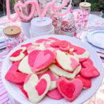 A plate of heart sugar cookies in four colors sits on a table decorated with pink accents, a neon “love” sign, sweets, and drinks, creating a festive and romantic atmosphere—these treats are delightful even with no icing.