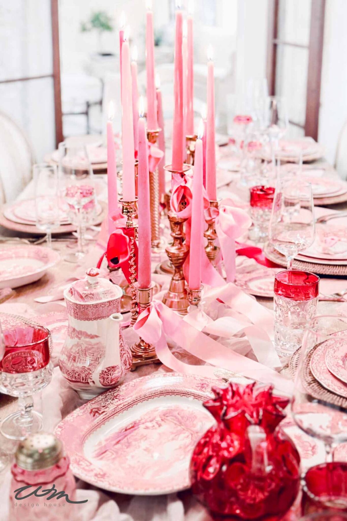 An elegant Valentine's Day tablescape set for a meal with pink and red dinnerware, crystal glasses, and gold candle holders holding tall pink candles, decorated with pink ribbons and gift bags. The overall atmosphere is romantic and festive.