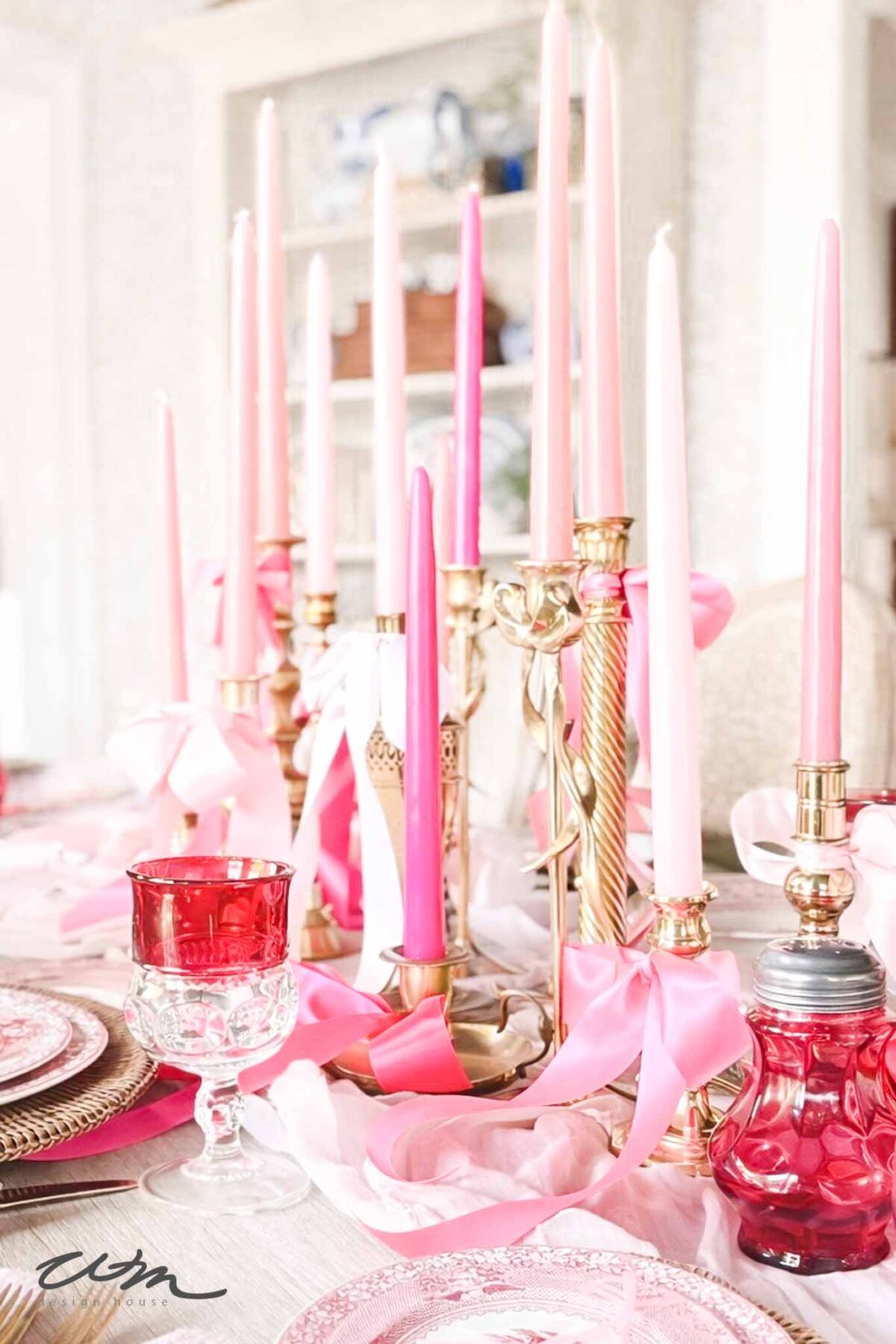 A romantic Valentine's Day tablescape with pink and red candles in gold holders, pink ribbons, a red glass goblet, pink napkins, and elegant plates creates a soft and festive atmosphere.