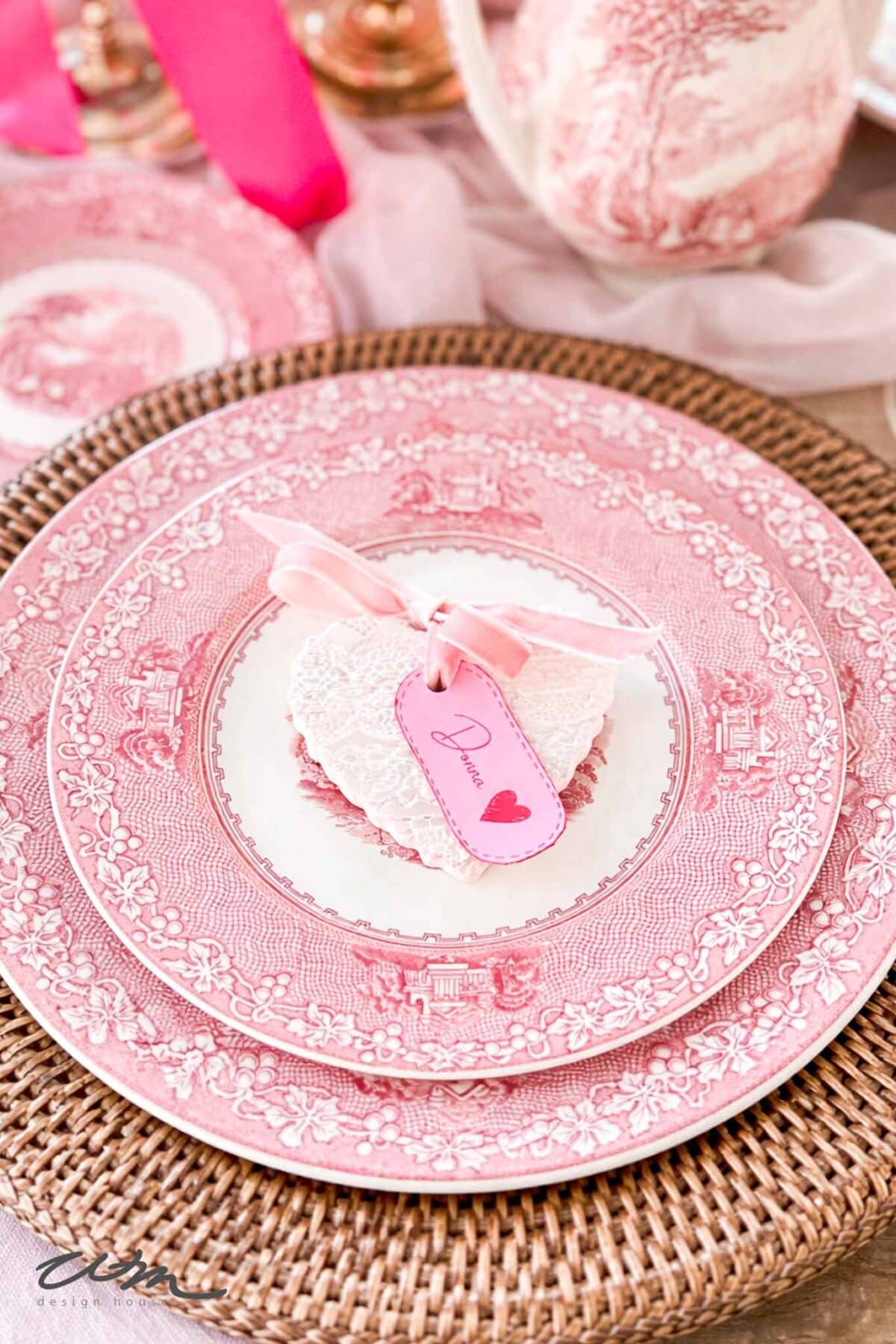 A Valentine's Day Tablescape featuring two pink floral plates on a woven charger, topped with a heart-shaped cookie and a pink ribbon tied to a tag adorned with a heart and the word "Darling.