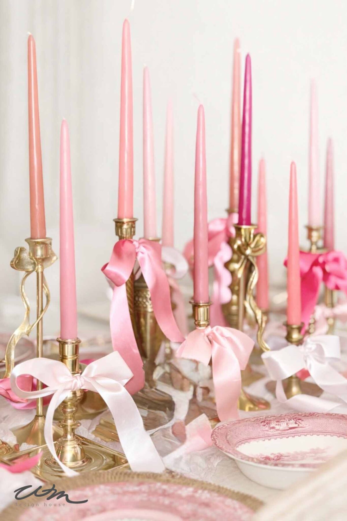 A Valentine's Day Tablescape features brass candle holders with pink taper candles in varying shades, each tied with delicate ribbons. Pink-patterned plates are elegantly arranged around the glowing centerpiece.