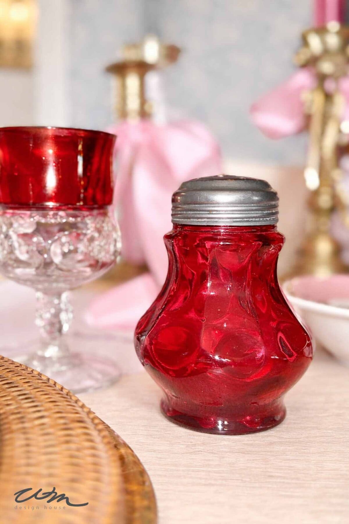 A vintage red glass salt shaker with a metal lid sits on a table next to a red and clear glass, with a wicker placemat partially visible in the foreground—perfect for a charming Valentine's Day tablescape.