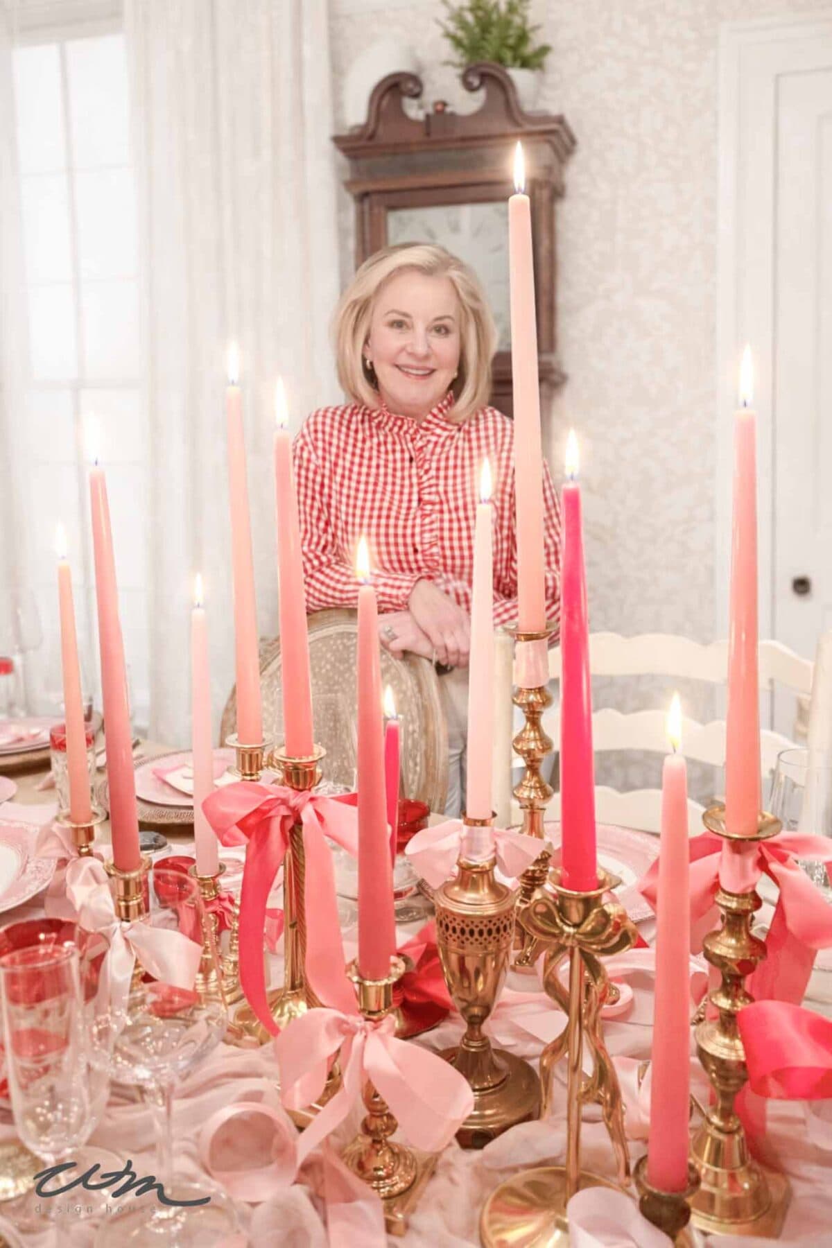 A woman in a red and white checkered blouse stands behind a Valentine's Day tablescape, featuring gold candle holders with pink candles and ribbons. The scene is bright and elegant, with a mirror and light walls in the background.