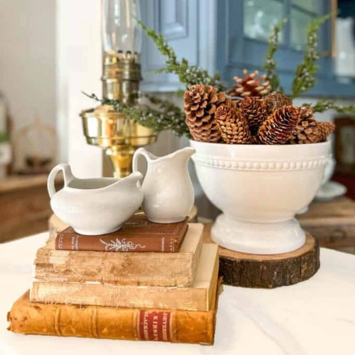 A stack of vintage books with two Vintage White Ironstone creamers on top sits next to a large white bowl filled with pinecones and greenery, all arranged on a white surface with a brass lamp in the background.