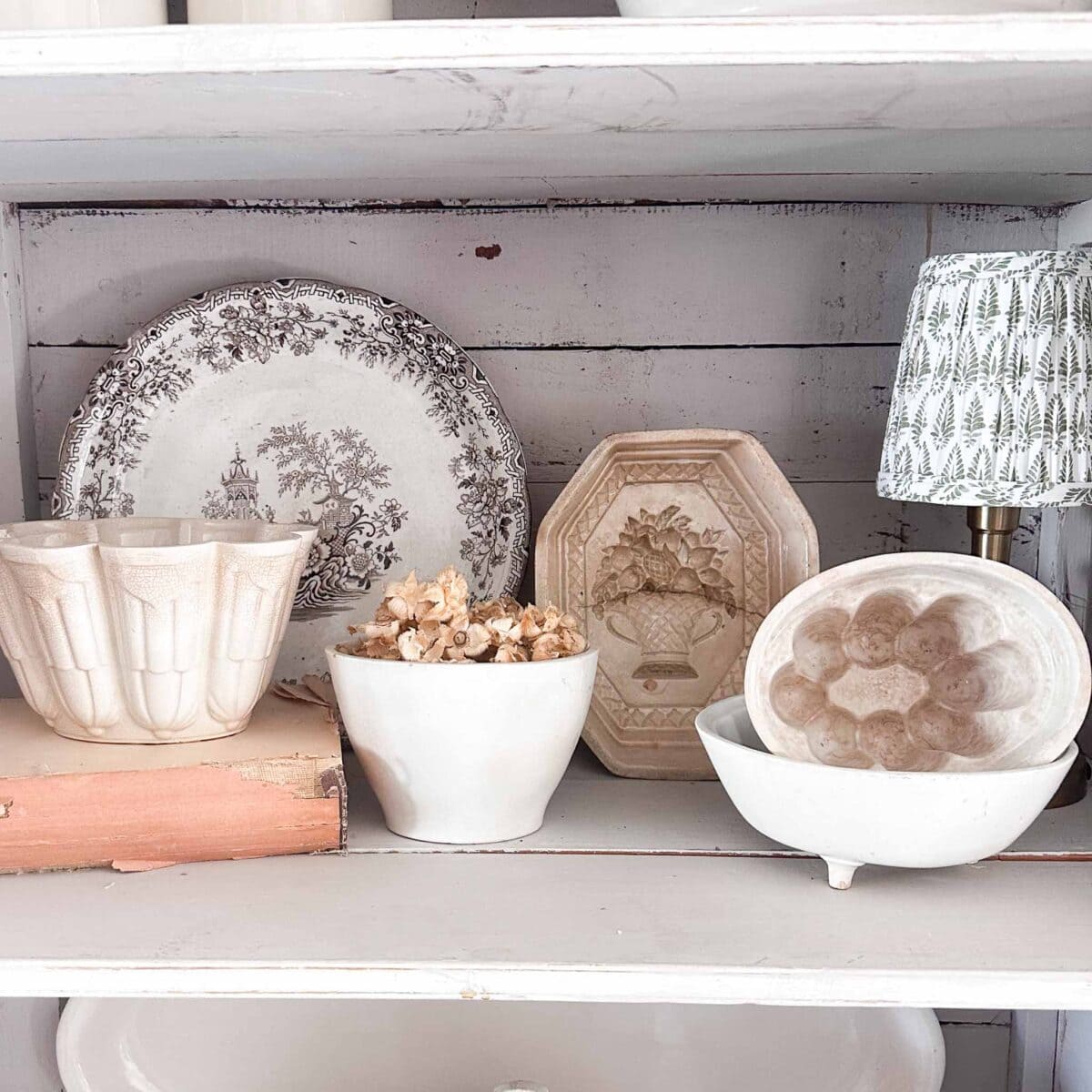 A white wooden shelf displays vintage ceramic bowls, a patterned plate, vintage white ironstone, a decorative mold, a bowl filled with dried flowers, and a small lamp with a green and white shade.