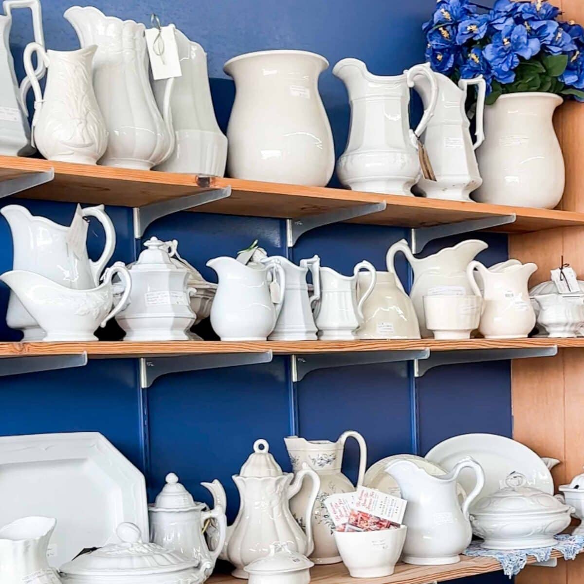 Shelves against a blue wall display an assortment of white ceramic pitchers, teapots, and serving dishes. A bouquet of blue flowers sits on the top right shelf, adding a pop of color to the arrangement.