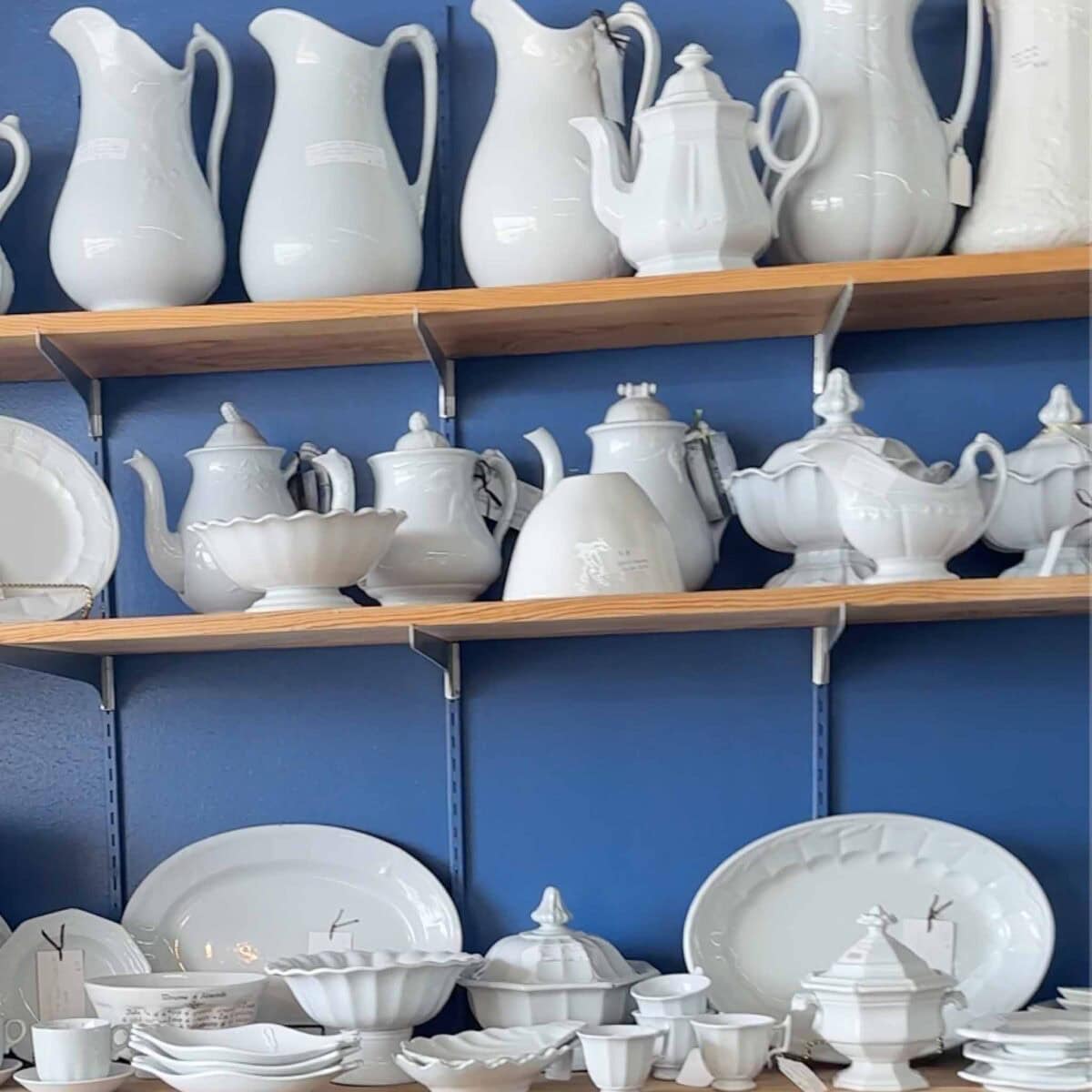Shelves mounted on a blue wall displaying various white ceramic dishes, pitchers, teapots, bowls, and plates arranged neatly in rows.