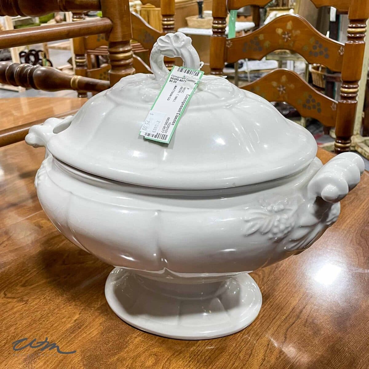 A vintage white ironstone soup tureen with ornate handles and a lid sits on a wooden table, with a price tag attached to the lid handle. Wooden chairs are visible in the background.