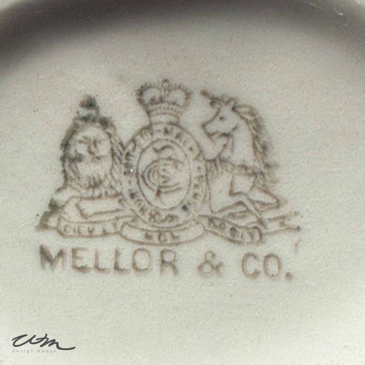 Faded stamp showing a lion and unicorn crest with a crown, encircling a shield, above the text "MELLOR & CO." on vintage white ironstone. A faint designer logo appears in the lower left corner.