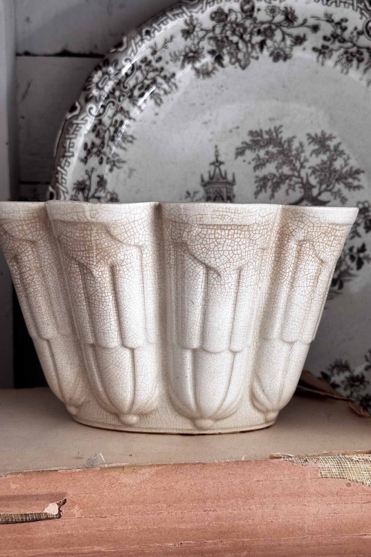 A vintage ceramic jelly mold with a crackled glaze sits in front of a decorative plate with a black pastoral design on a wooden shelf.