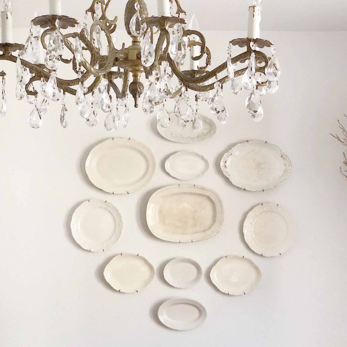 A vintage chandelier with crystal pendants hangs above a white wall decorated with a symmetrical arrangement of various Vintage White Ironstone and ceramic plates.