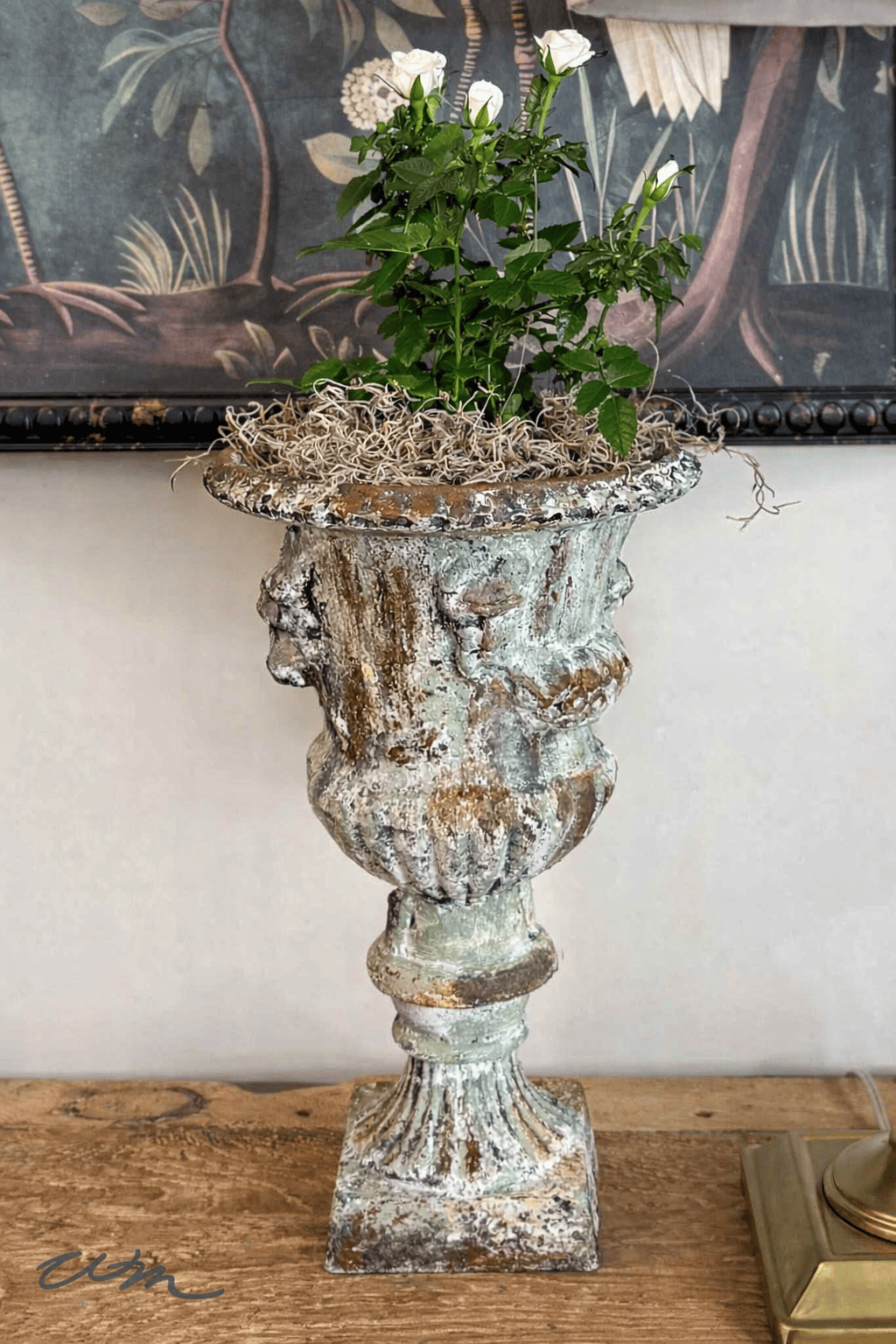 A weathered, ornate urn planter brings vintage charm to your spring entry table decor, holding a small rose bush with white blooms and moss. The planter rests on a wooden surface before a decorative wall art background.