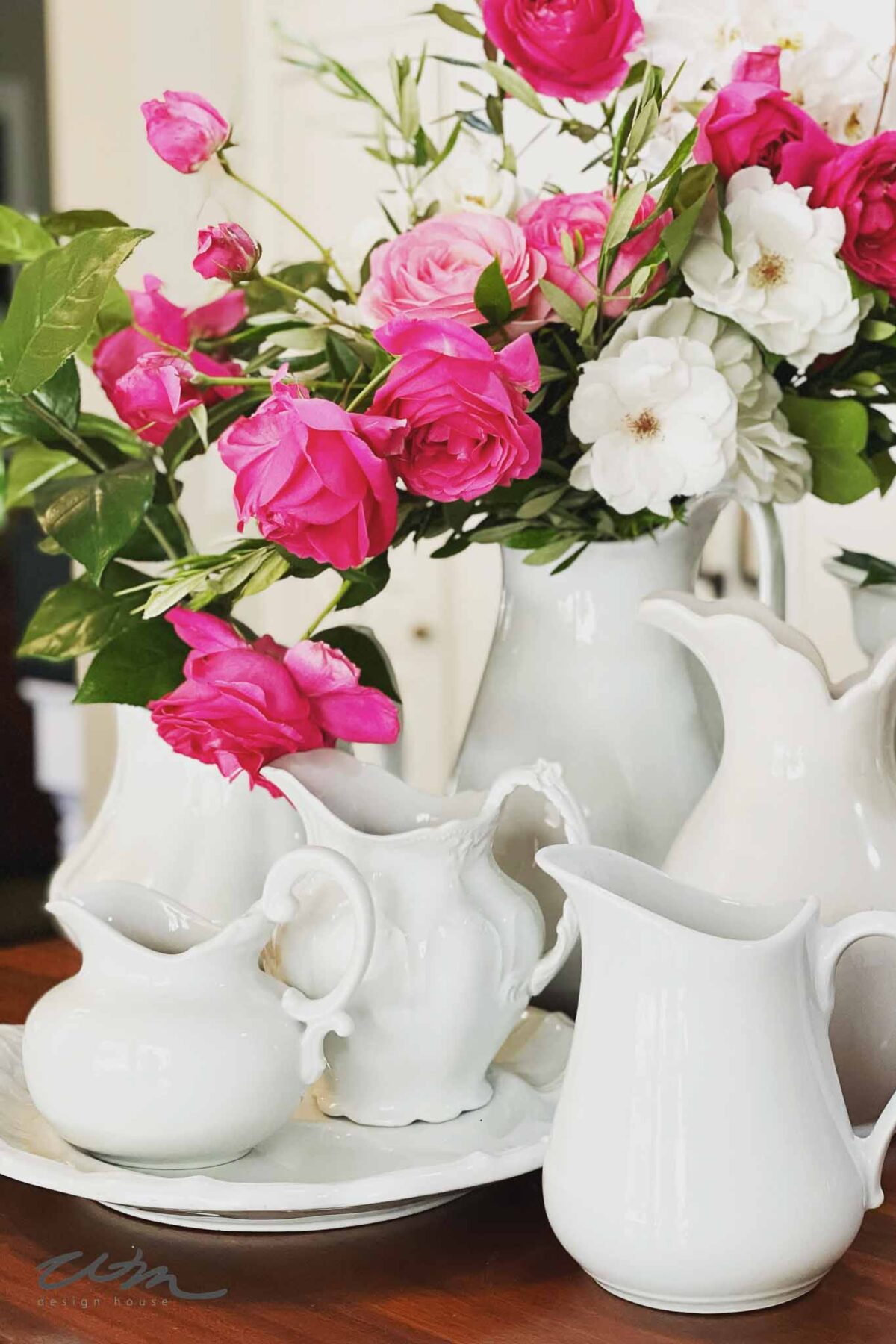A bouquet of pink and white roses in a white ceramic pitcher, surrounded by several white ceramic pitchers and a plate, all arranged on a wooden surface.