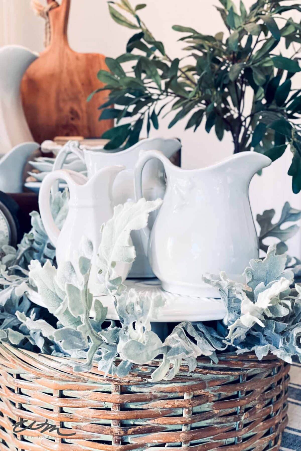 Two white ceramic pitchers are displayed in a woven basket, surrounded by soft, dusty green foliage. A wooden board and leafy plant are in the background, creating a cozy, rustic arrangement.