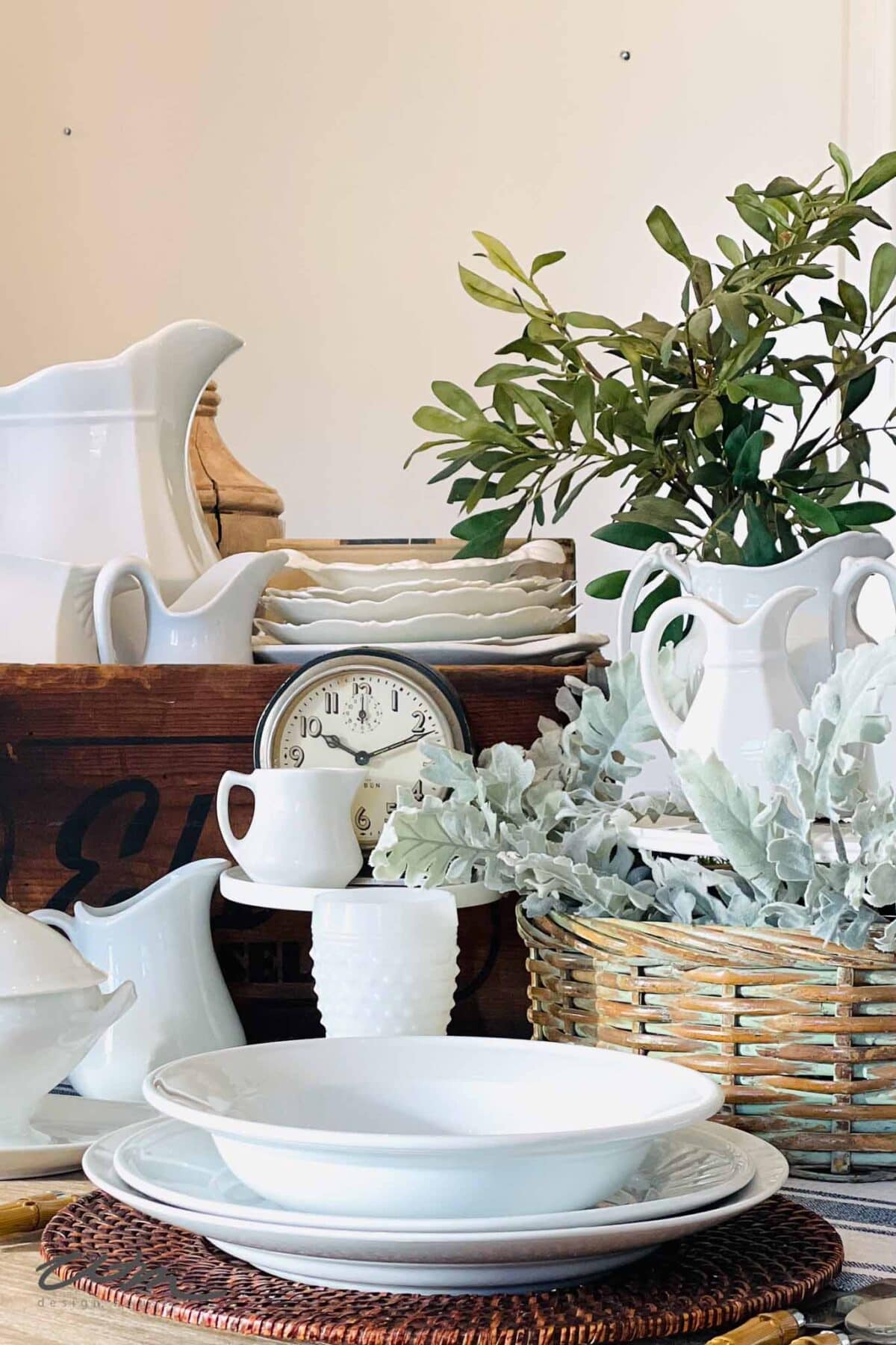 A collection of white ceramic dishes and pitchers arranged on a table with woven trays, a vintage clock, and green and silver foliage in baskets, creating a rustic, farmhouse-style display.