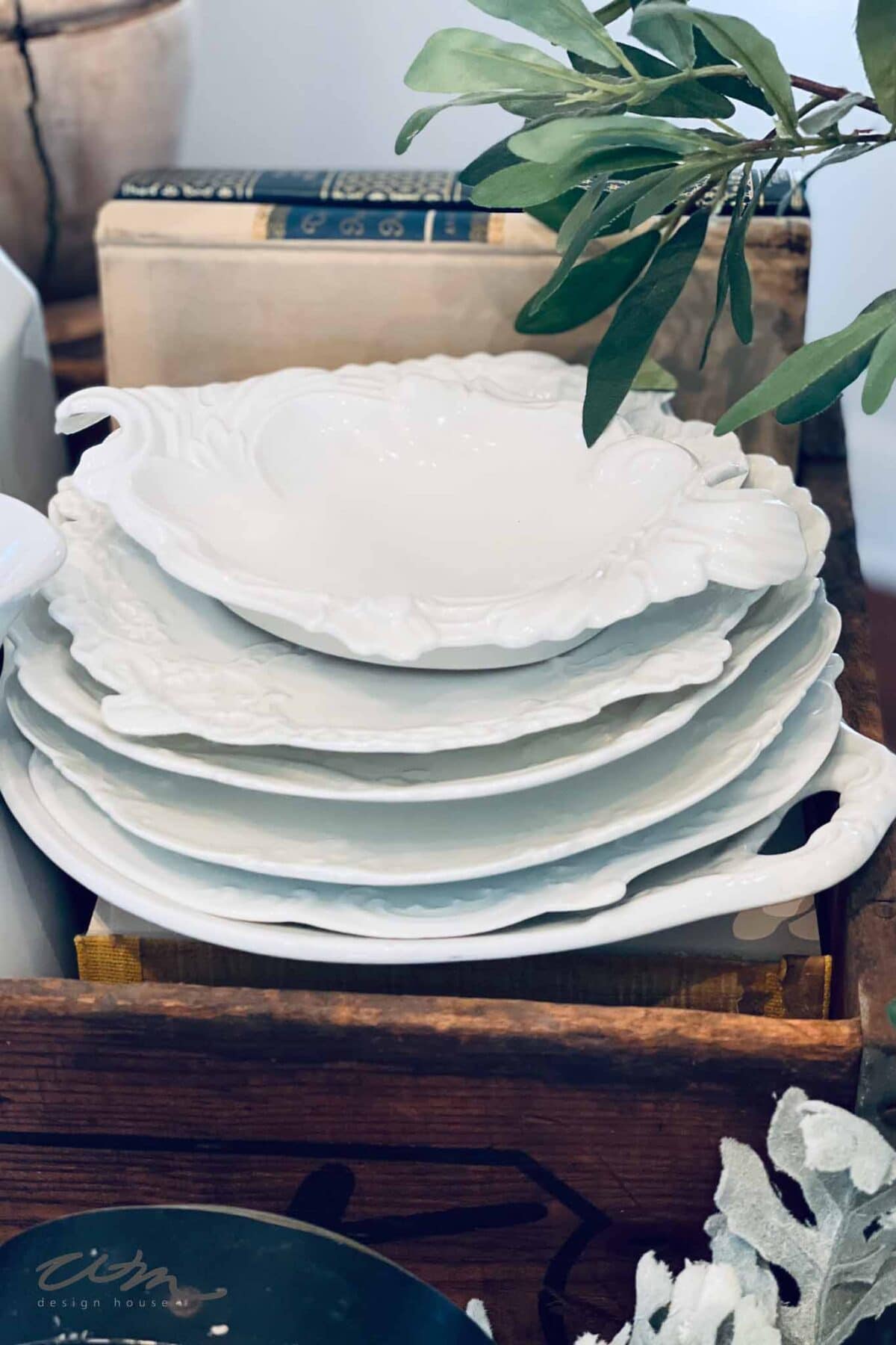 A stack of ornate white ceramic plates sits in a wooden crate, surrounded by green leaves and old books, creating a vintage, rustic arrangement.