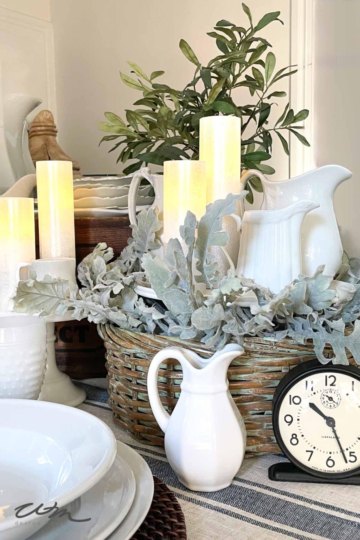 A tabletop arrangement features white ceramic pitchers, glowing LED candles, leafy greenery in a wicker basket, a black and white clock showing 3:55, and stacked white dishes on a striped cloth.