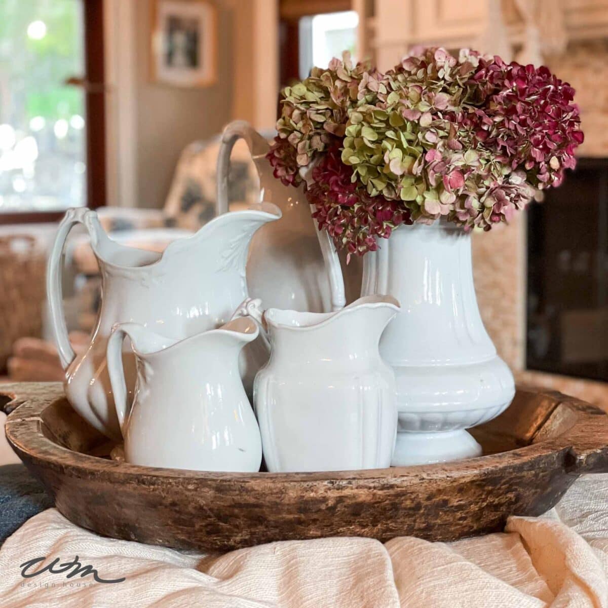 A rustic wooden tray holds four white ceramic pitchers and vases, one filled with pink and green hydrangeas, set atop a textured cloth on a table in a cozy, softly lit living space.