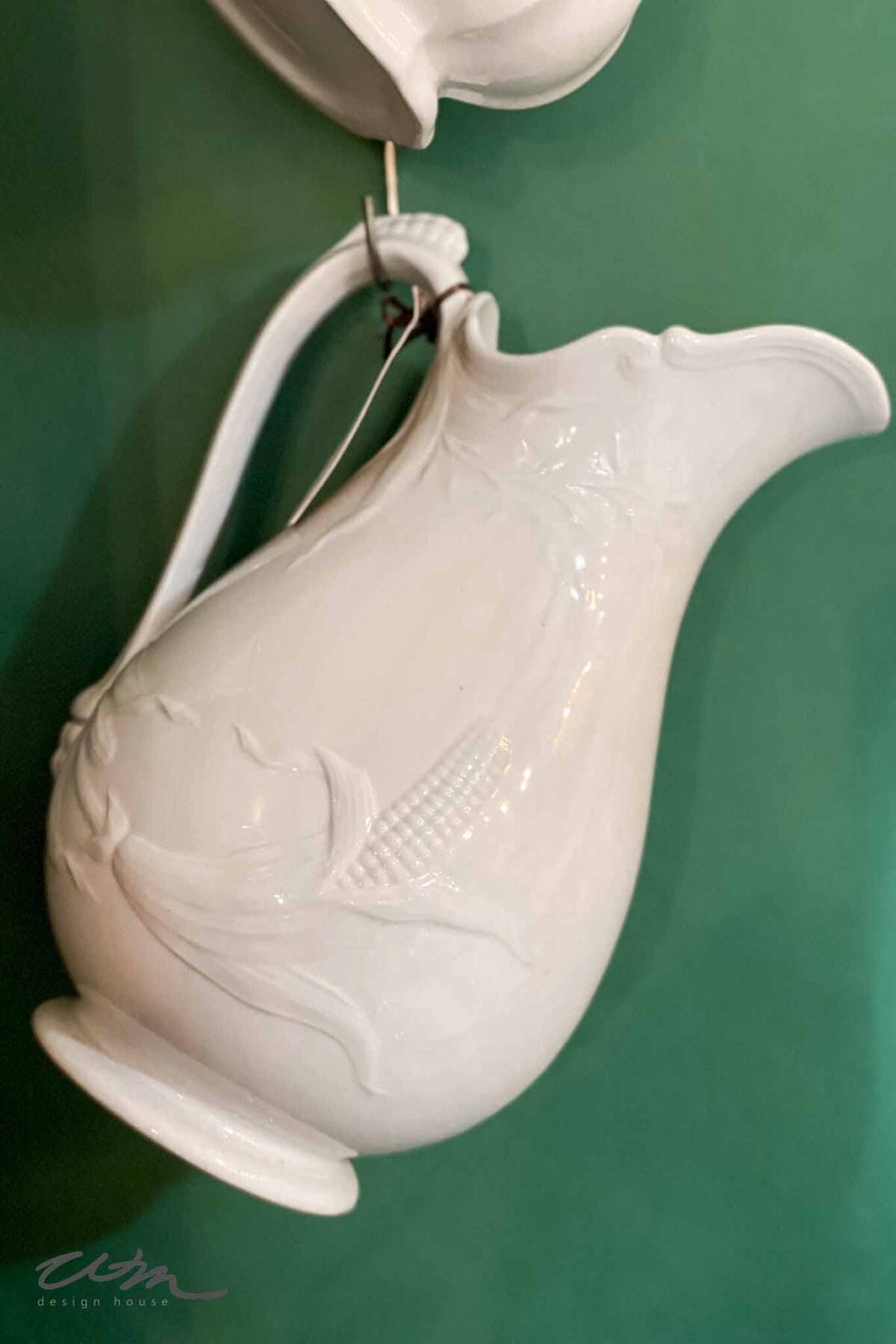A white ceramic pitcher with ornate detailing hangs against a green background. The handle is curved, and subtle embossed patterns decorate the side of the pitcher.