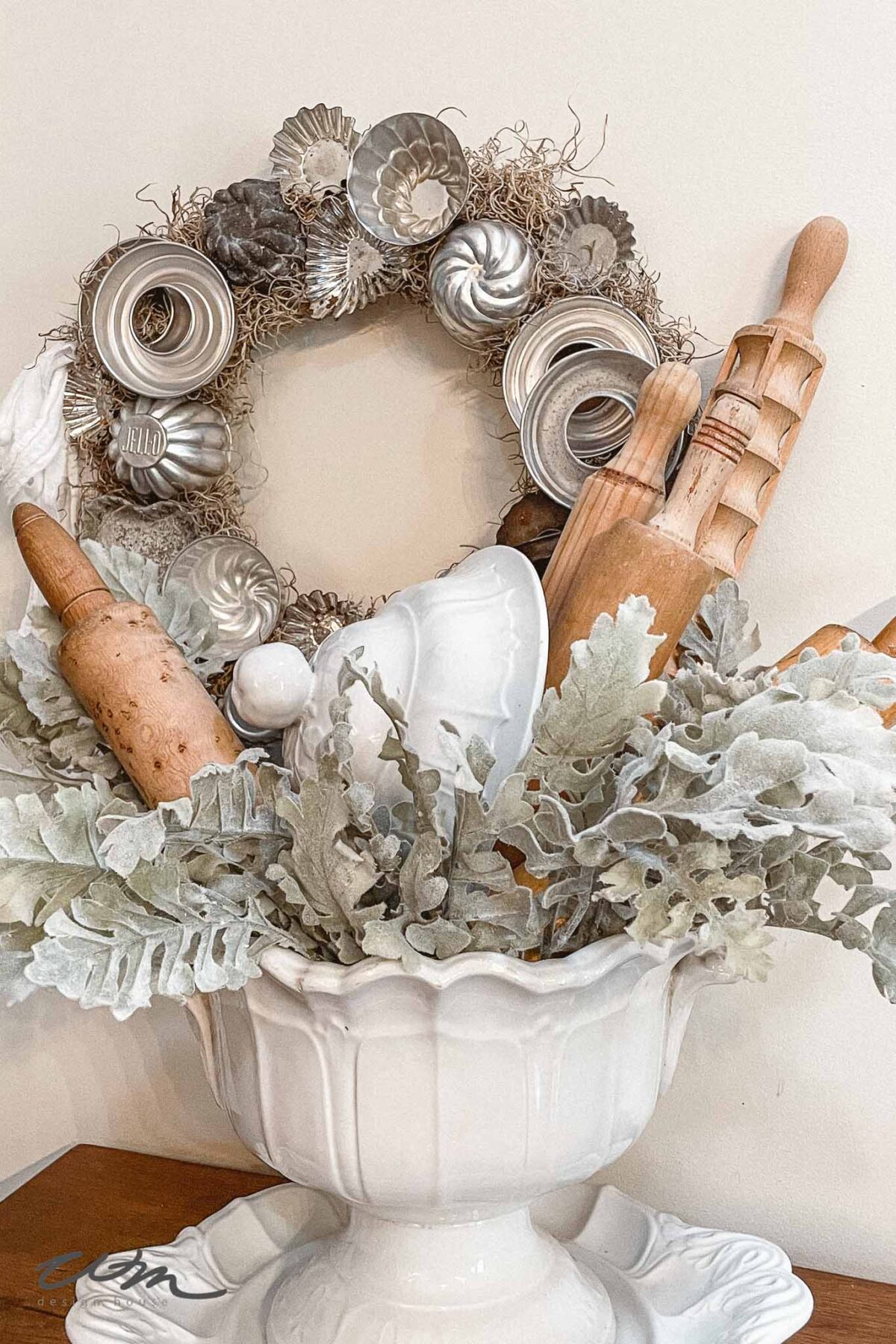 A white ceramic bowl holds rolling pins and baking tools among silvery faux leaves. Behind it, a wreath made of vintage metal tart molds hangs on the wall, creating a rustic and cozy kitchen decor display.