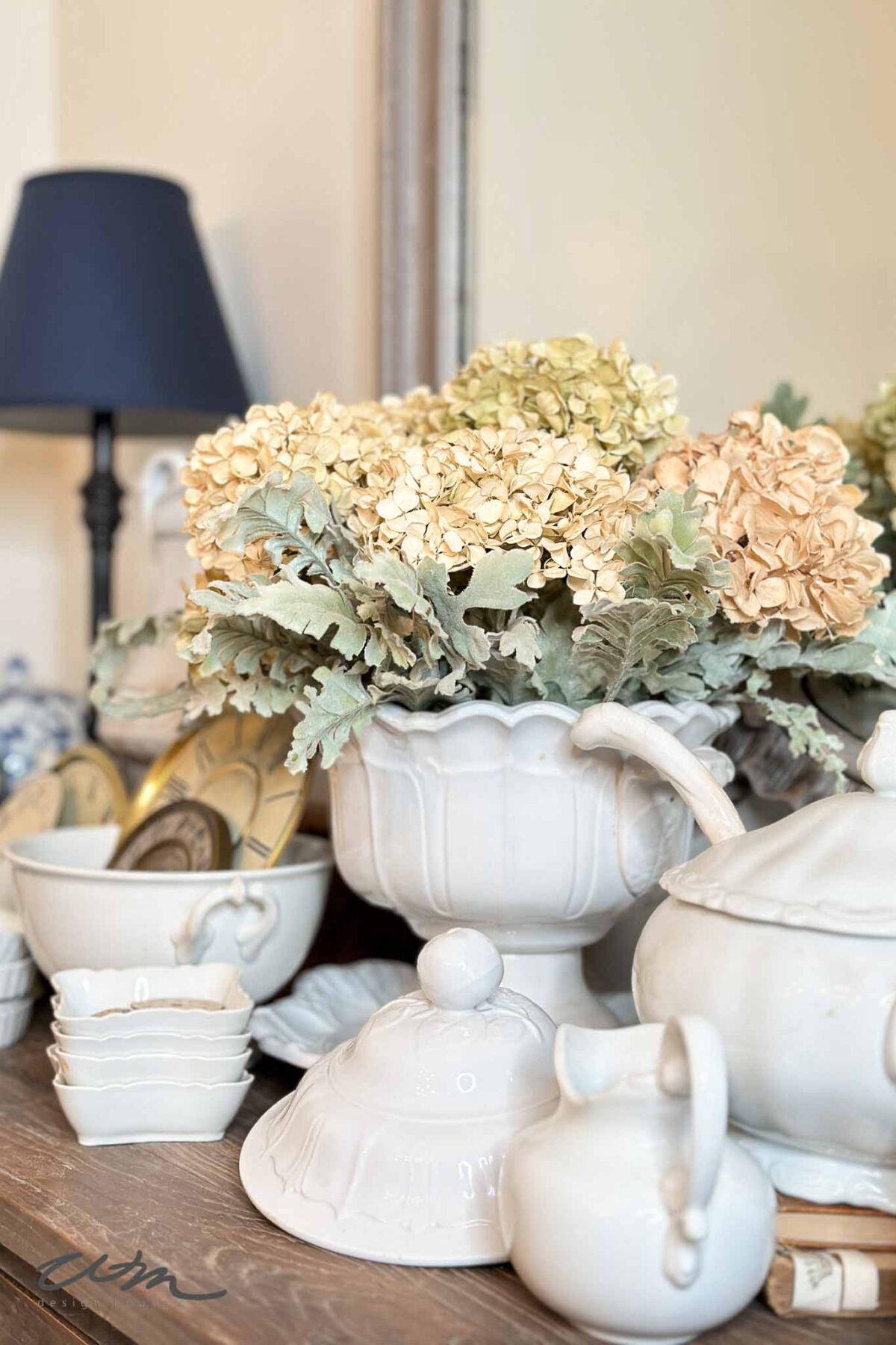 A decorative arrangement of cream-colored dried flowers in a white ceramic vase, surrounded by various white ceramic dishes, bowls, and a teapot on a wooden surface, with a black lamp in the background.
