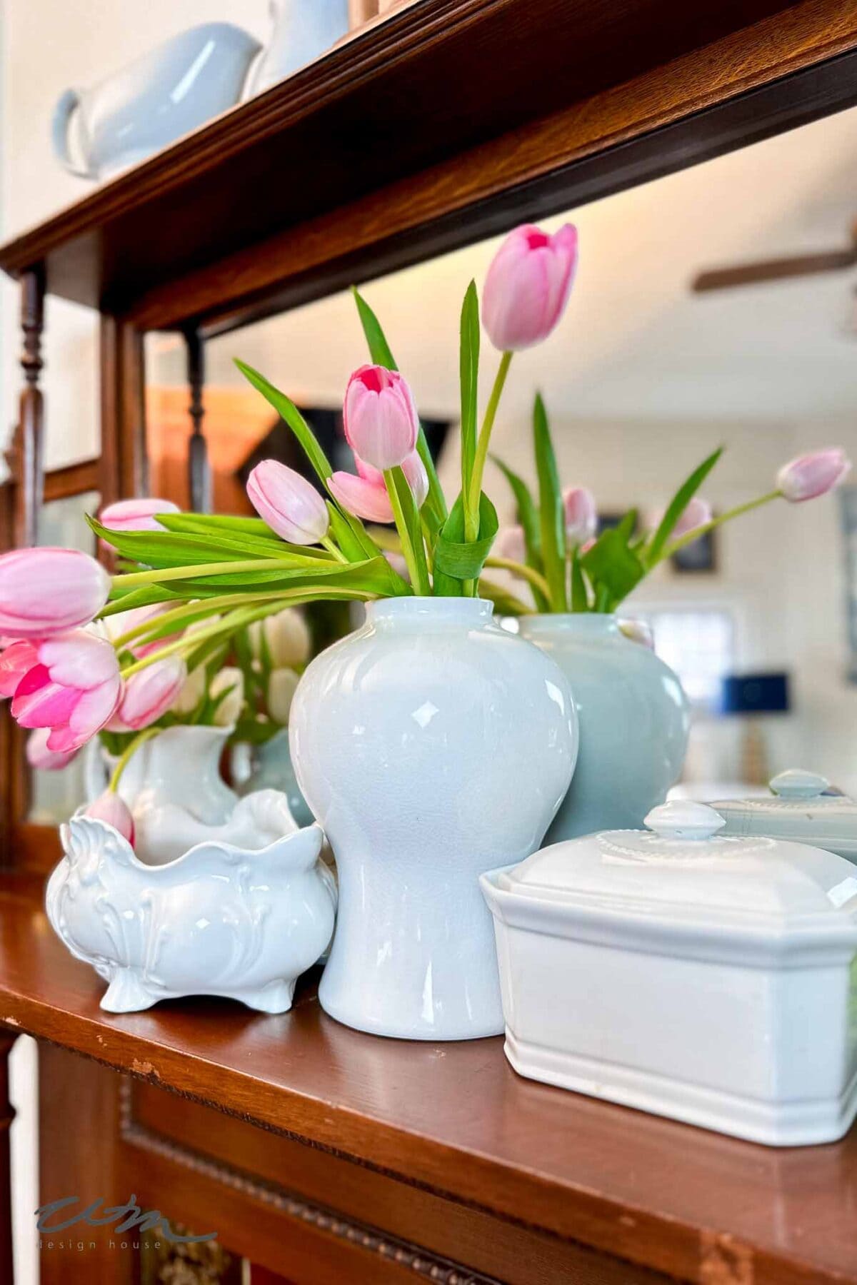 A white ceramic vase with pink tulips sits on a wooden shelf beside other white porcelain dishes, all reflected in a mirror behind them. The background shows a softly lit room.