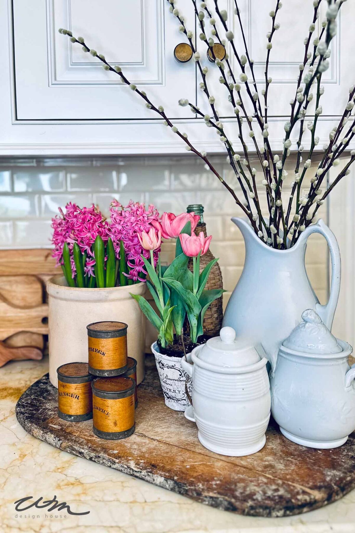 A kitchen countertop decorated with pink hyacinths, tulips, and pussy willow branches in vases, vintage spice jars, and white ceramic containers on a rustic wooden tray.
