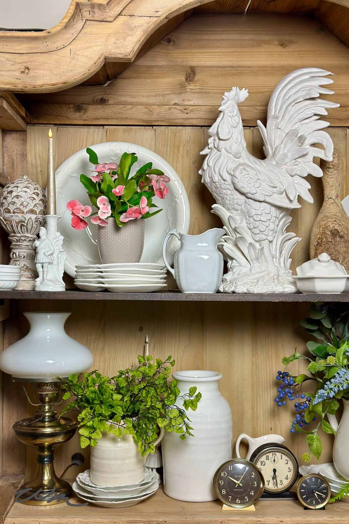 A wooden shelf displays white ceramic dishes, a tall white rooster figurine, flower arrangements, an antique lamp, two vintage clocks, and decorative greenery. The background is rustic wood paneling.