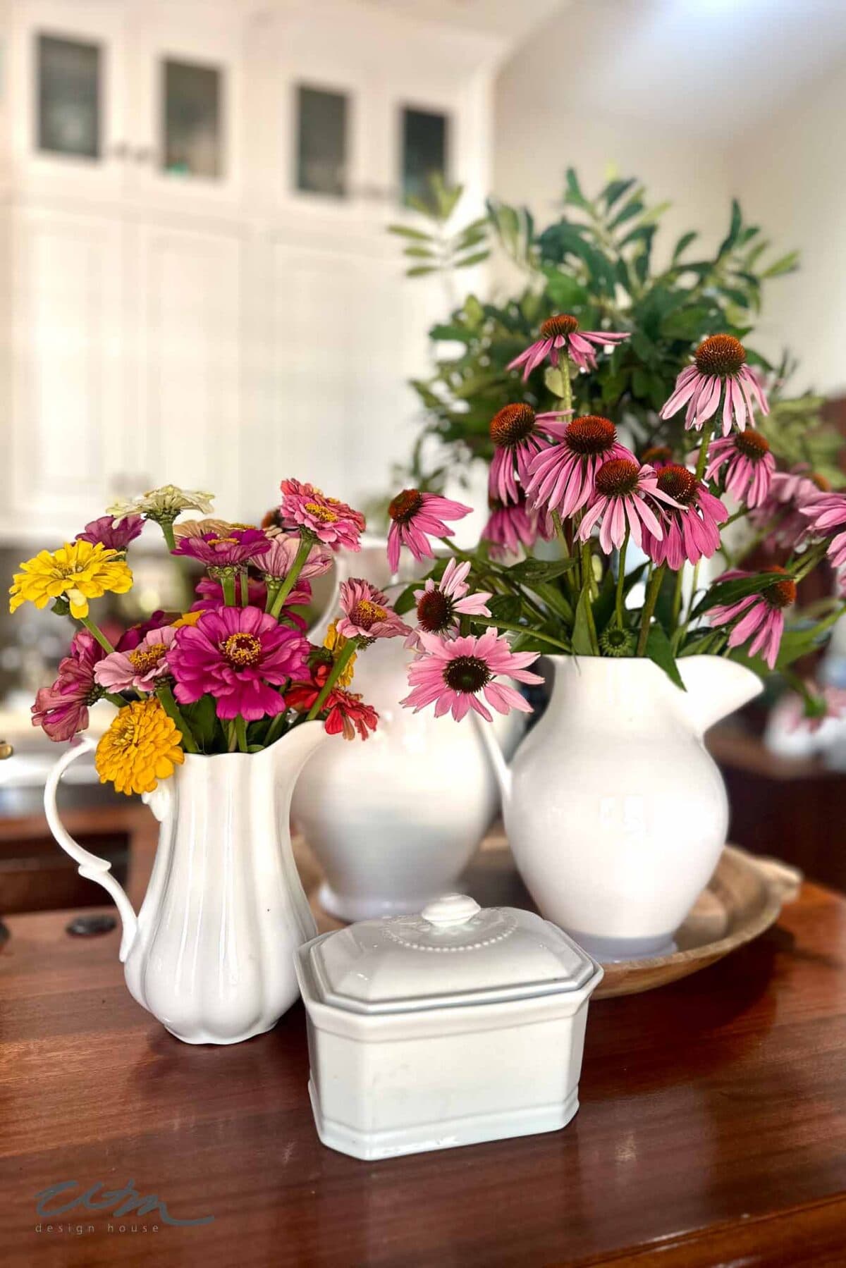 A wooden table displays white ceramic pitchers filled with colorful flowers, including pink coneflowers and yellow zinnias. A white ceramic box with a lid sits in front. Blurred white cabinets are in the background.