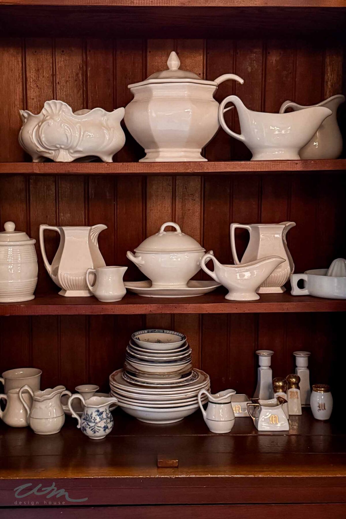 A wooden cupboard displays neatly arranged white ceramic dishes, bowls, pitchers, cups, and tureens, along with a few small blue-and-white and gold-accented items on the shelves.