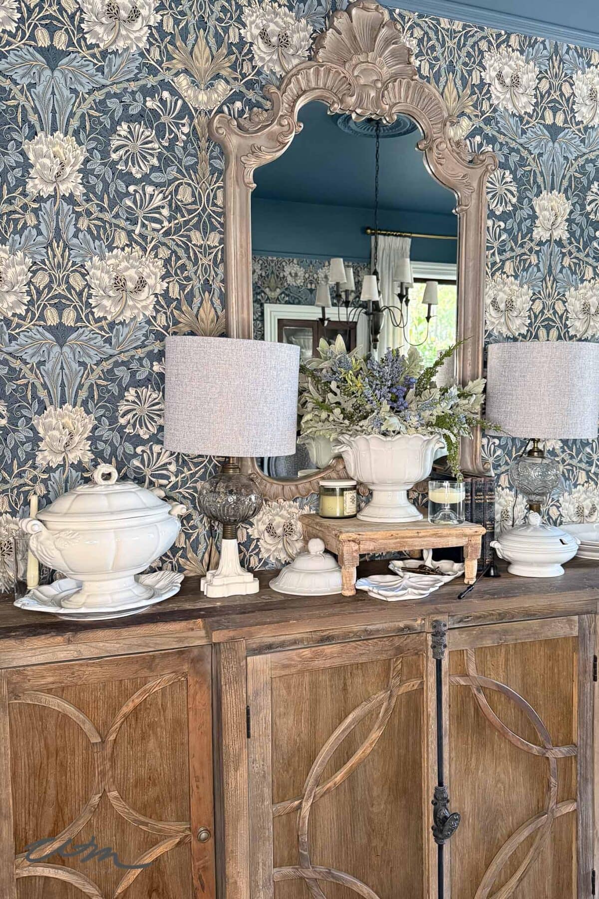 A wooden sideboard with ornate cabinet doors holds white ceramic dishes, two table lamps with gray shades, and a flower arrangement. An ornate mirror hangs above, reflecting a chandelier and part of the room. Blue floral wallpaper in the background.