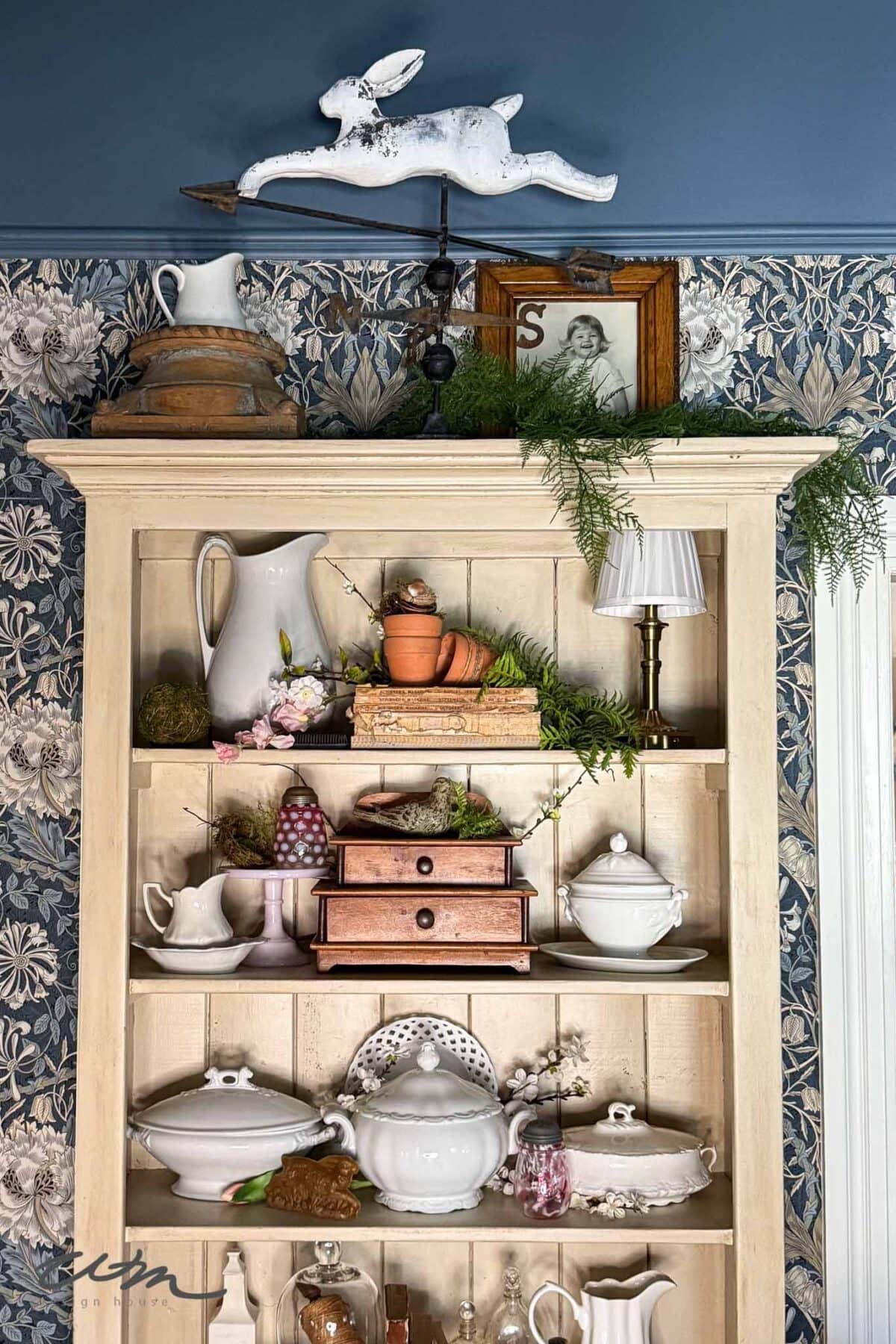 A cream-colored hutch displays white ceramic dishes, pitchers, and teapots, with some copper items and spring decor. A white rabbit weather vane sits on top, alongside greenery, a lamp, and framed art. Blue floral wallpaper is in the background.