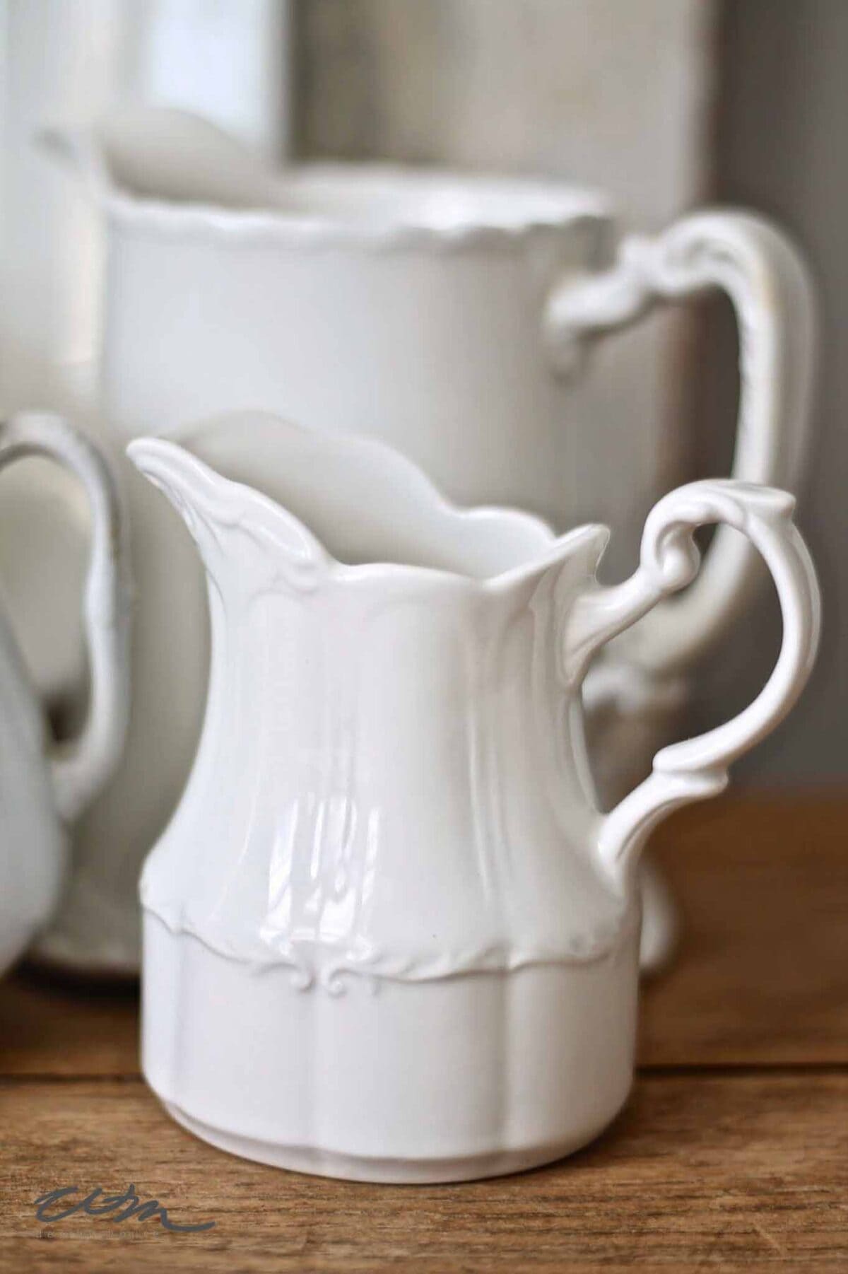 A small, ornate white ceramic pitcher with a decorative handle sits on a wooden surface in front of a larger, similarly styled white pitcher. Both have scalloped edges and elegant curves.