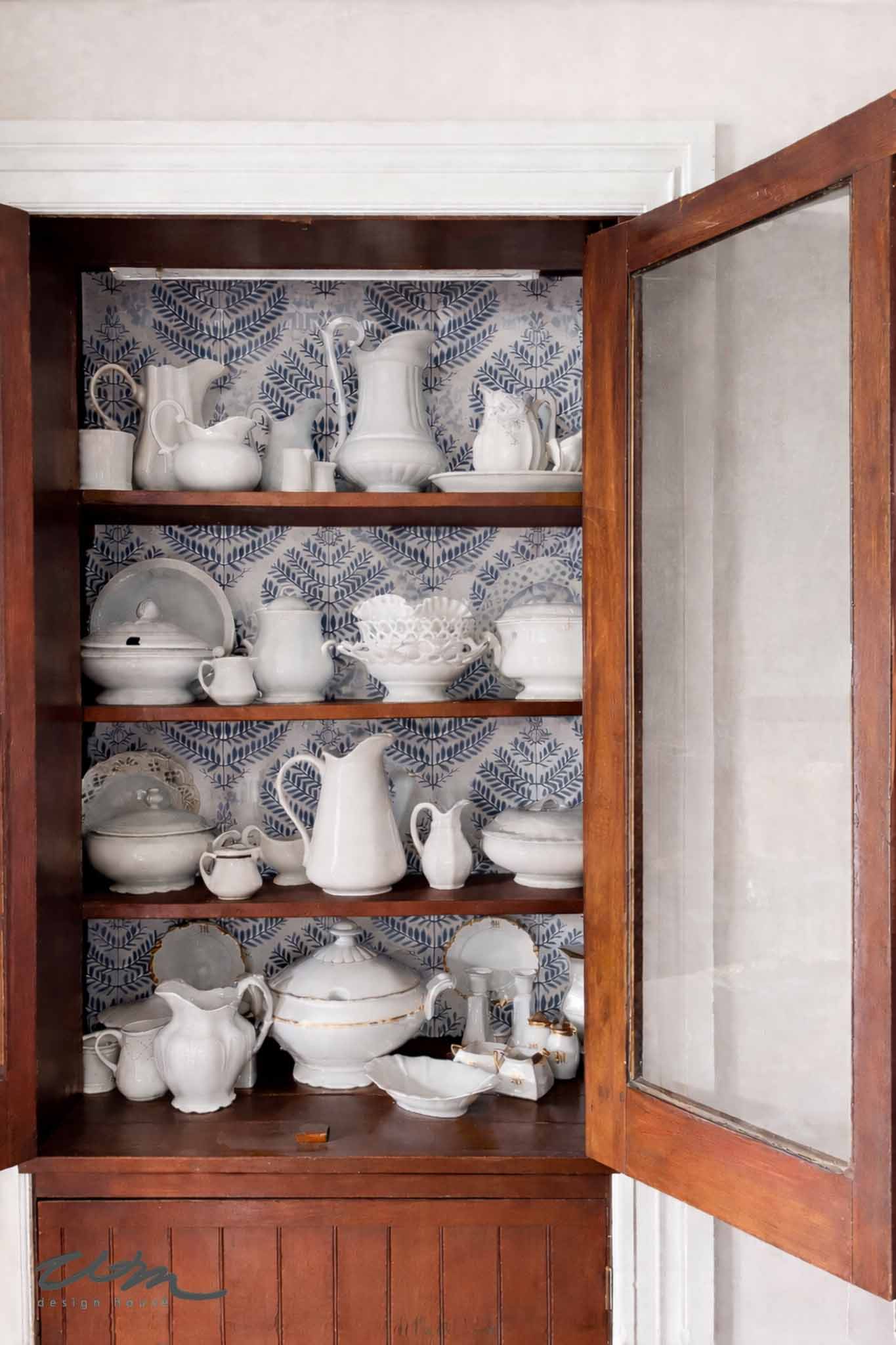 A wooden cabinet with glass doors displays shelves filled with vintage white ironstone china and pitchers, set against a blue-and-white leaf-patterned wallpaper backdrop.