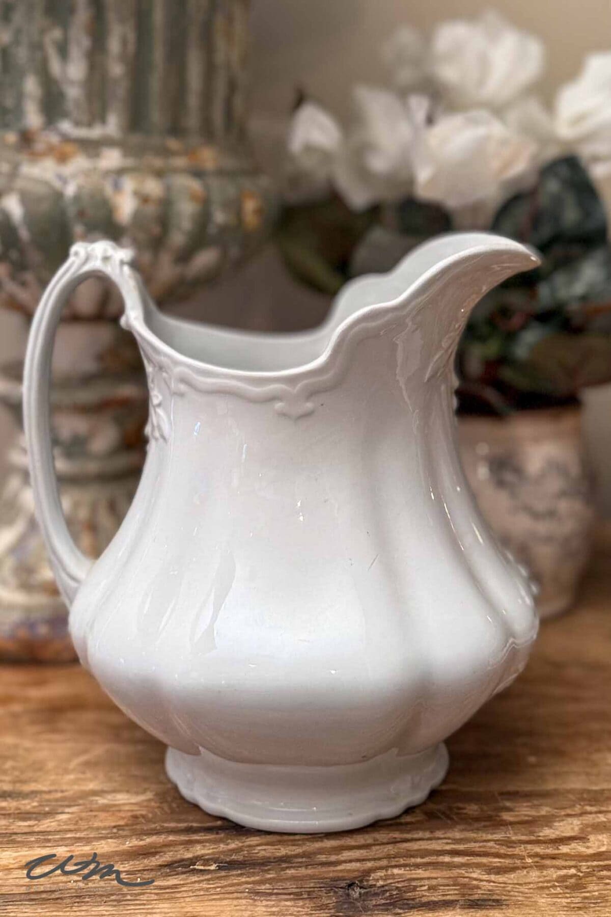 A white, ornate ceramic pitcher with a curved handle and scalloped rim sits on a wooden surface, adding vintage charm to spring entry table decor. Blurred white flowers and a decorative vase create an inviting background.