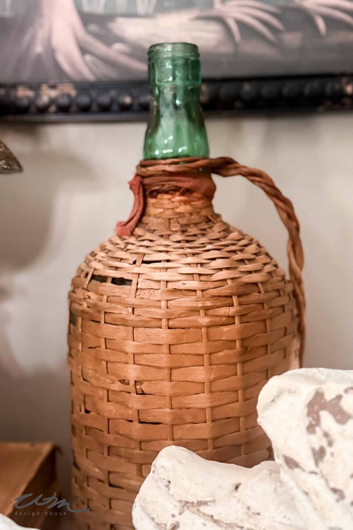 A green glass bottle, encased in a woven wicker basket with a handle, exudes vintage charm as it sits on a surface next to light-colored rocks. The blurred background enhances its appeal—perfect for Spring entry table decor.