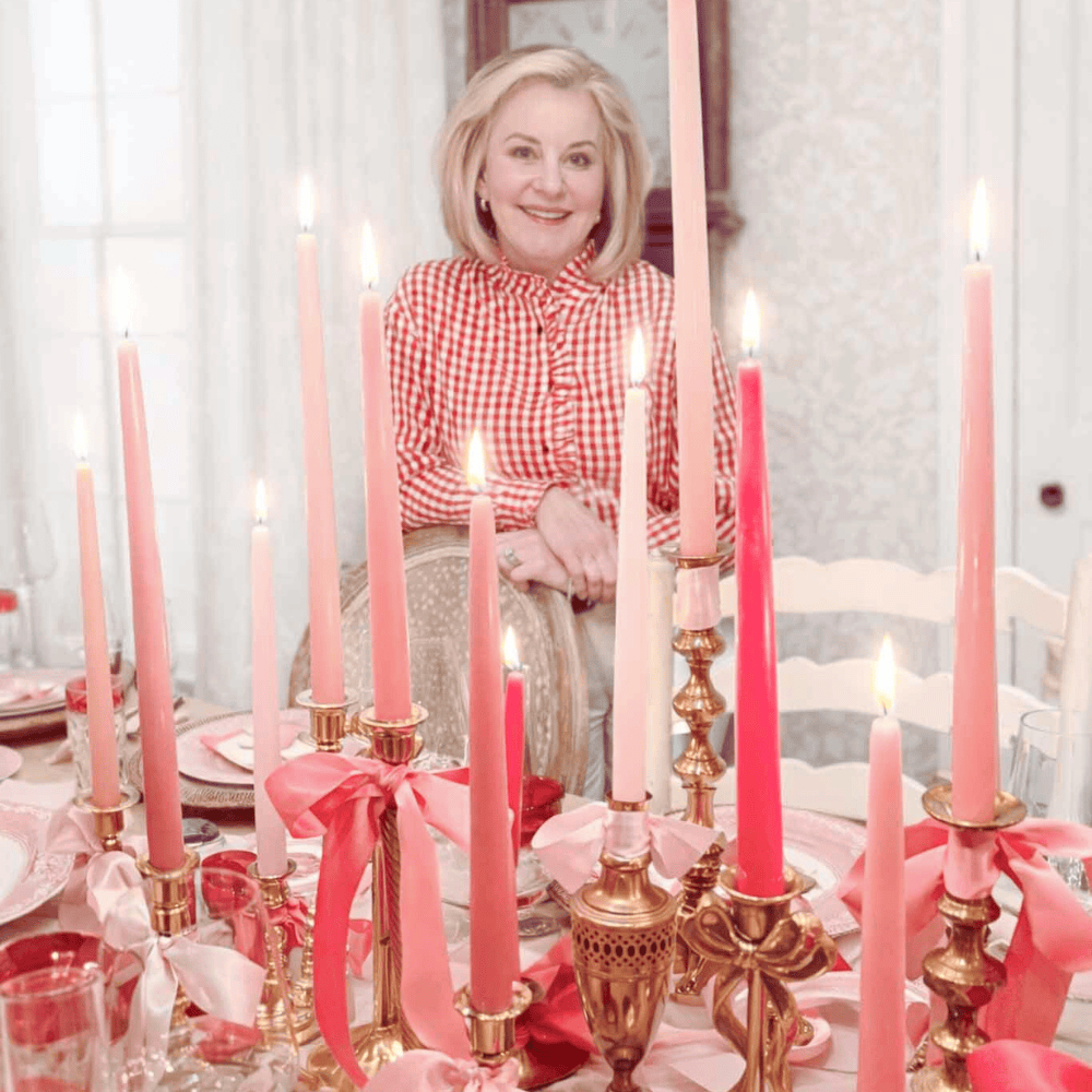 A woman in a red and white gingham blouse smiles while sitting at a decorated table featuring a Valentine's Day tablescape with tall pink, red, and white candles in gold holders, adorned with ribbons for a festive atmosphere.