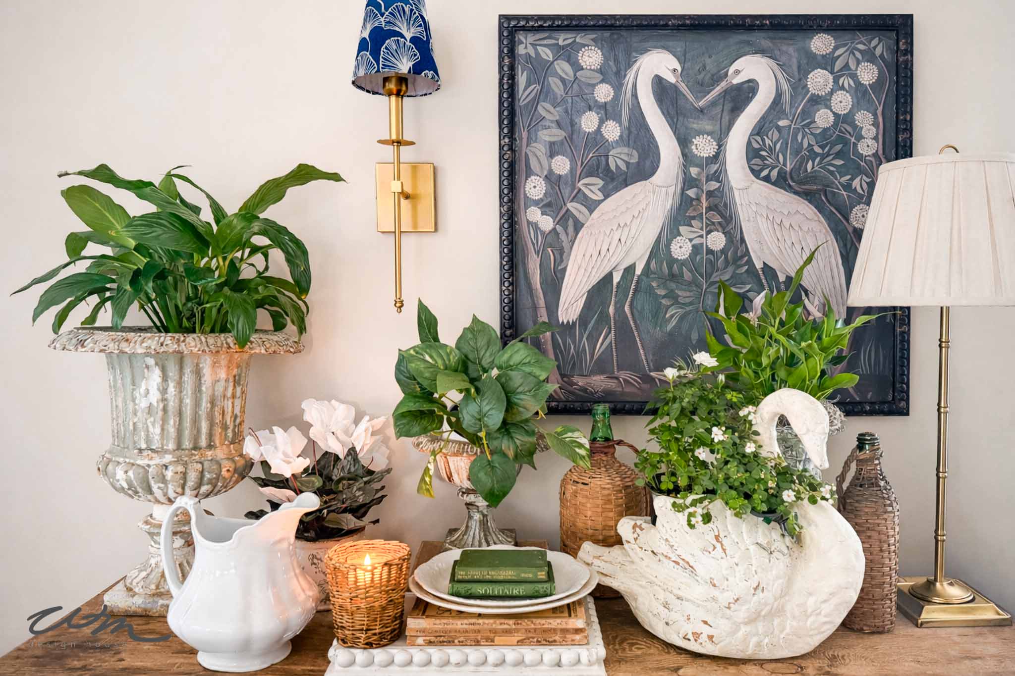 A decorative entry table decor features potted plants, a white swan planter, a lit candle, books, a white pitcher, and a lamp, with framed artwork of two white cranes hanging on the wall behind.