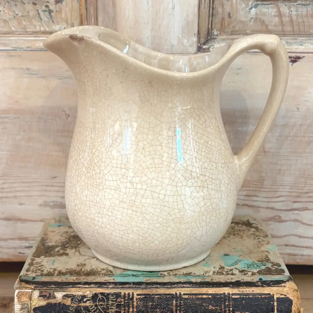 A vintage white ironstone cream-colored ceramic pitcher with a crackled glaze sits atop two weathered, stacked books with faded covers, set against a distressed wooden background.