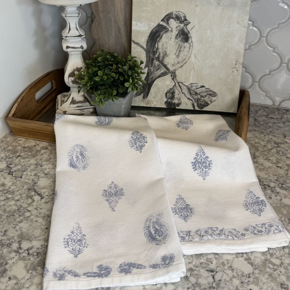 A kitchen countertop display with a wooden tray holding a small potted plant, a linen towel with blue patterns, and a framed bird sketch leaning against the wall.