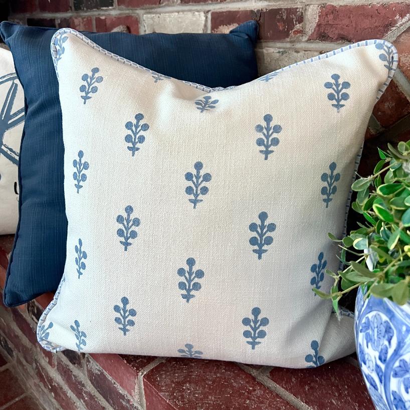 A decorative white pillow with a blue leaf pattern rests on a brick bench, accompanied by a dark blue pillow and a potted green plant in a blue and white ceramic pot.