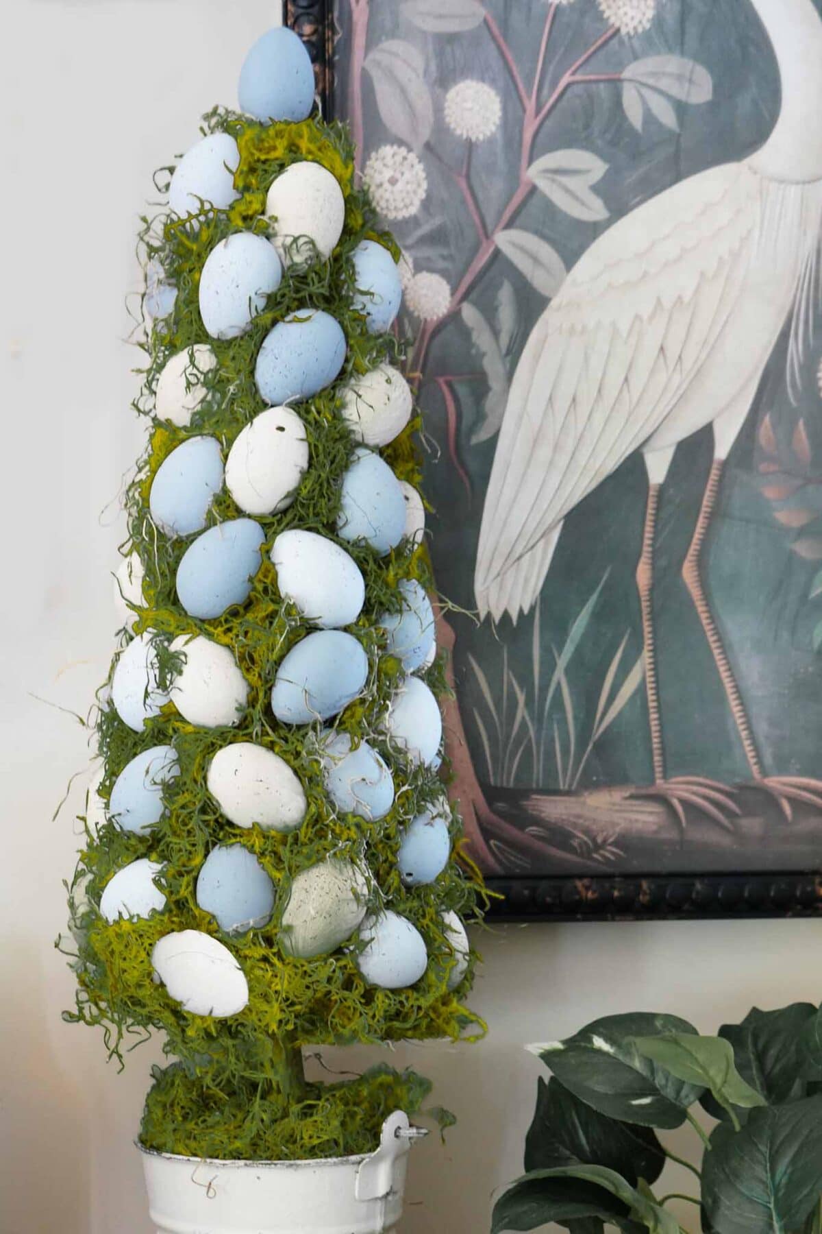 A decorative tree-shaped arrangement with blue and white eggs and green moss, set in a white pot—perfect for vintage Easter decorating ideas. A leafy plant and framed bird artwork add charming touches in the background.