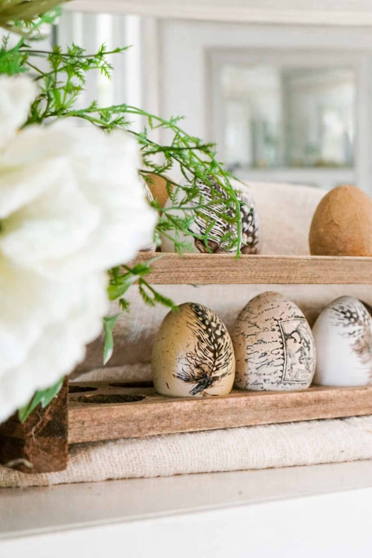 Decorative eggs with black and brown botanical designs are displayed on a wooden tray next to greenery and white flowers, offering charming spring decor inspiration and vintage Easter decorating ideas.
