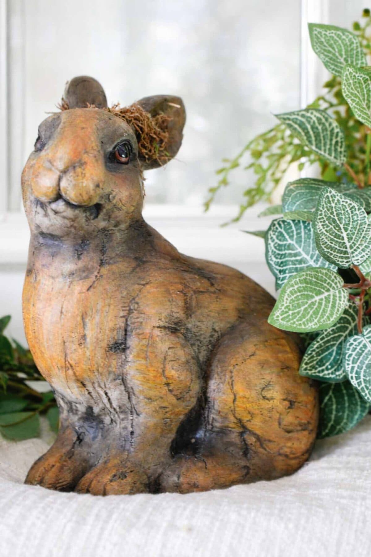 A realistic brown rabbit statue sits on a light surface, surrounded by green leafy plants, perfect for vintage Easter decorating, with a window blurred in the background.