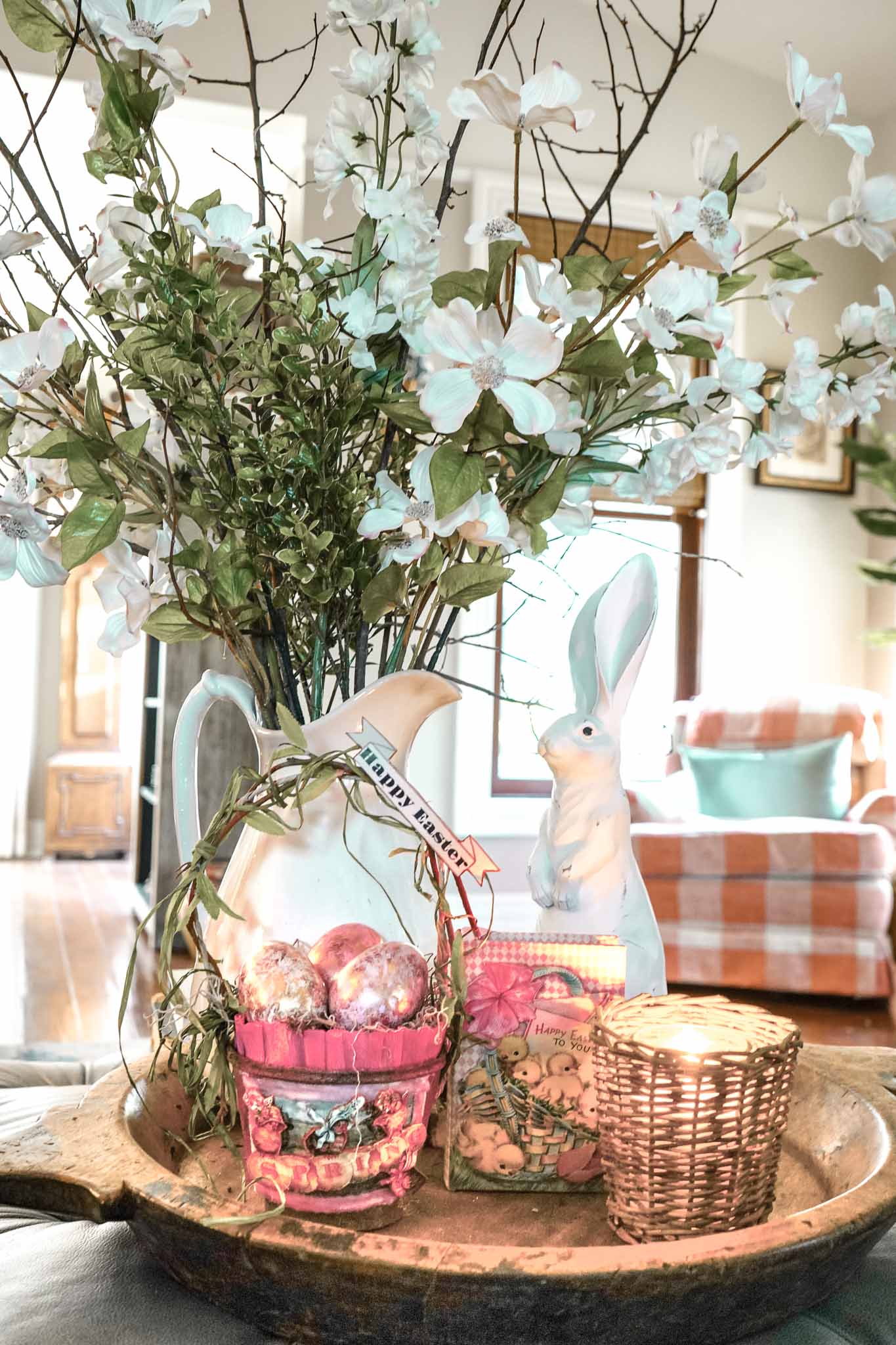 A decorative tray showcases simple Easter decor: a vase with white flowers, a ceramic bunny, a pink basket of shiny eggs, and a candle in a wicker holder—perfect inspiration for vintage Easter decorating in your cozy living room.